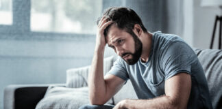 Unpleasant pain. Sad unhappy handsome man sitting on the sofa and holding his forehead while having headache
