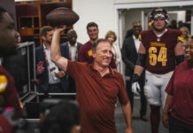 Washington Commanders’ Josh Harris on Making Sports a ‘Shared Community Experience’ Photo of a smiling man holding a football in the air. He is surrounded by players from the Washington Commanders. All are wearing maroon shirts.