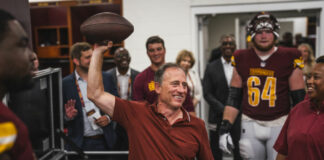 Photo of a smiling man holding a football in the air. He is surrounded by players from the Washington Commanders. All are wearing maroon shirts.