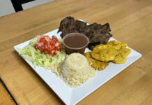 Authentic Salvadoran Cuisine in Montgomery County Photo of a plate of food on a wooden table with lettuce and pico de gallo, white rice, meat and some yellow patties around a clear plastic cup of brown sauce.