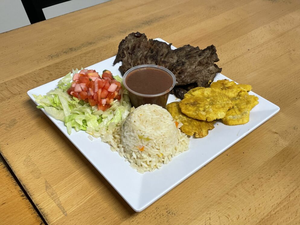 Photo of a plate of food on a wooden table with lettuce and pico de gallo, white rice, meat and some yellow patties around a clear plastic cup of brown sauce.