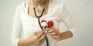 Stock photo of a woman wearing a stethoscope and holding it up to a pretend red heart. She is wearing a gold ring and a bracelet.
