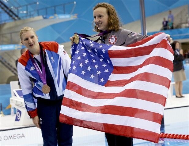 Photo of Katie Ledecky smiling with a gold medal around her neck. She is holding up an American flag.