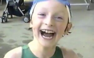 Photo of a six-year-old girl with a huge grin on her face looking at the camera. She is wearing a swim cap with goggles on her head.