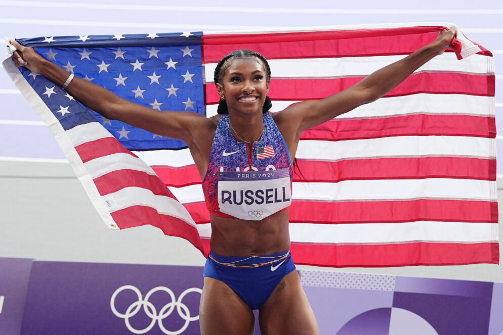 Photo of a woman wearing the U.S. track and field uniform with her arms outstretched holding a large American flag. Her bib, pinned to the front of her shirt, says "Russell."