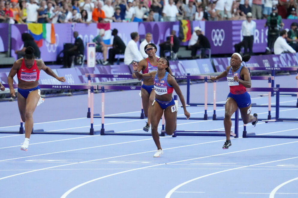 Photo of women competing in a hurdling event on a blue track in front of hundreds of spectators.