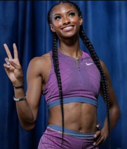 Photo of a young woman with long dark hair in two braids past her waist. She is wearing a matching Nike workout set, purple spandex with blue stripes. She is holding up a peace sign with one hand and smiling at the camera. Her six pack of abs is visible.