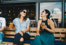 Stock photo of two women wearing sunglasses and sitting on a bench laughing together. They are each holding a glass of beer.