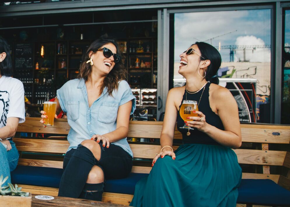 Stock photo of two women wearing sunglasses and sitting on a bench laughing together. They are each holding a glass of beer.