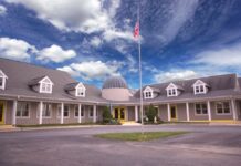 Photo of the exterior of a gray school building with two wings. At the center is the entrance with a tall flagpole with the American flag out front.