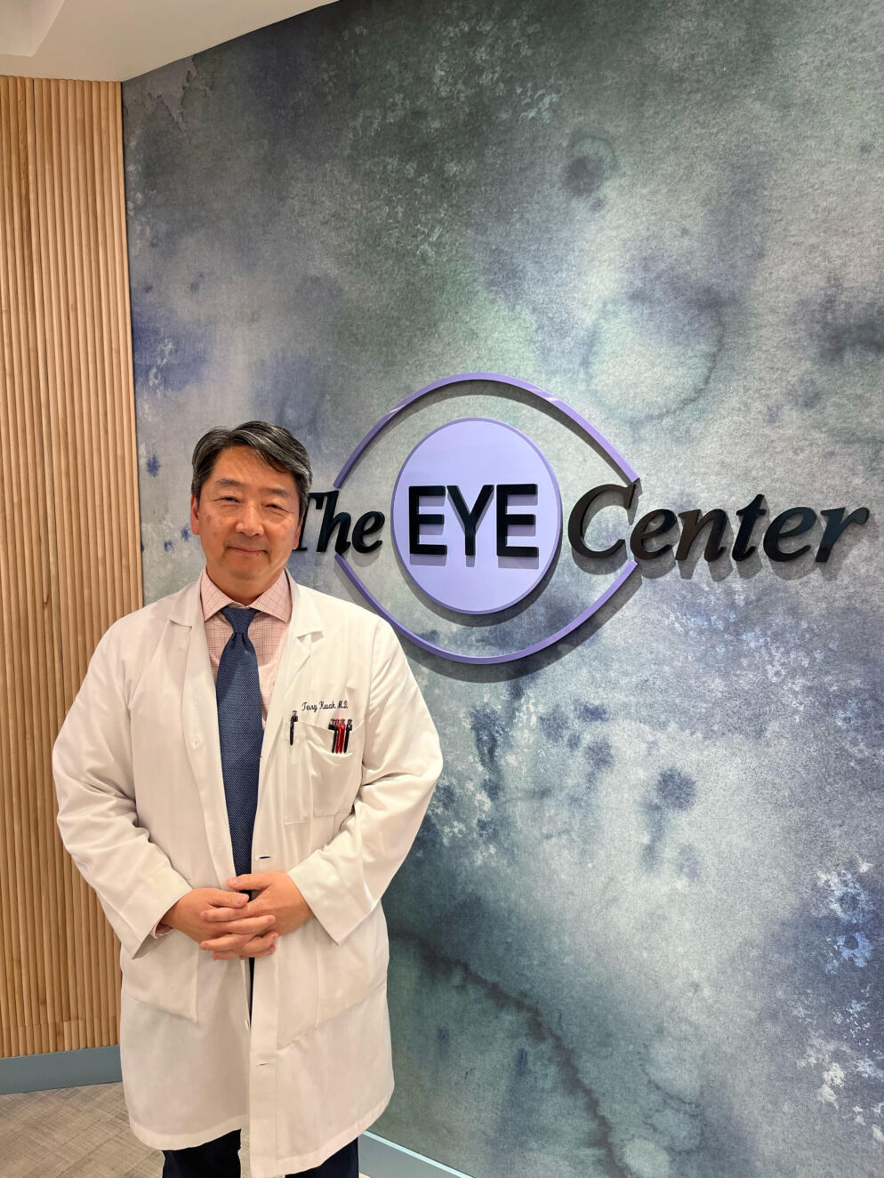 Photo of a man with short dark hair. He is wearing a white doctor's coat with a light pink shirt and blue tie, posing in front of a blue wall with The Eye Center logo.