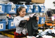 Photo of a woman holding up a pair of black shorts as she sorts through clothes in a warehouse. Behind her are about 10 blue bins of clothes.