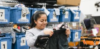 Photo of a woman holding up a pair of black shorts as she sorts through clothes in a warehouse. Behind her are about 10 blue bins of clothes.