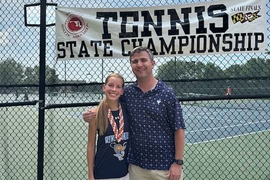Photo of a man with his arm around a young woman with a medal around her neck. They are standing outside a tennis court below a banner that reads "Tennis state championship."