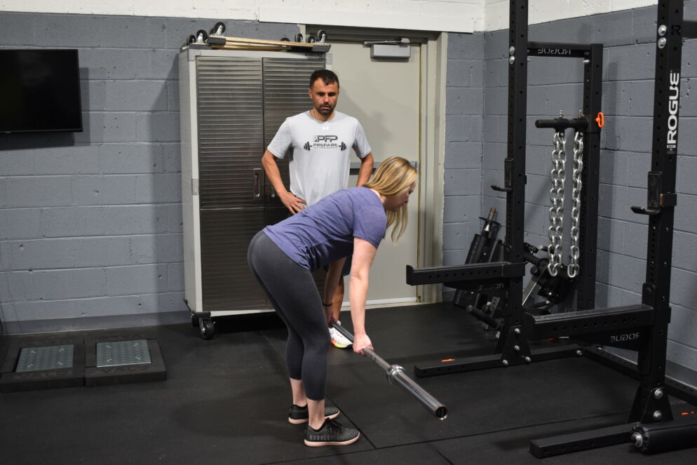 Photo of a blonde woman deadlifting a bar in a fitness studio as a coach looks on in the background.