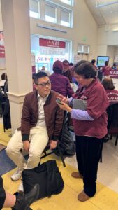 Photo of a young Vietnamese man with short dark hair and glasses helping an older Vietnamese woman use her cell phone in a classroom-like setting.