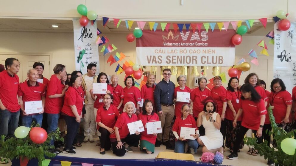 Group photo of about two dozen Vietnamese older adults posing together wearing red T-shirts. A banner above them reads "VAS Senior Club."