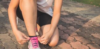 Stock photo of a woman lacing up her running shoes, gray sneakers with pink laces. She is wearing running clothes: a gray T-shirt and black shorts.
