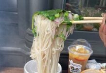 Photo of a bowl of rice noodles and green herbs in broth. A person's hand holding chopsticks lifts noodles out of the bowl.