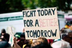 Stock photo of a protester holding up a cardboard sign that reads "Fight today for a better tomorrow" in hand-drawn letters.