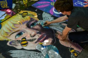 Stock photo of a man with short dark hair drawing with chalk on the ground. He is drawing a realistic portrait of a blonde person surrounded by flying bees.