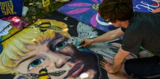 Stock photo of a man with short dark hair drawing with chalk on the ground. He is drawing a realistic portrait of a blonde person surrounded by flying bees.