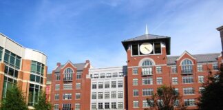 Photo of a town center. There is a large brick building with a clock tower, trees and city benches where people are sitting.