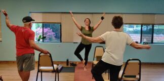 Photo of a woman doing a tree pose: standing on one leg with the other bent, and two arms raised in the air, in front of a yoga class.