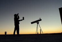 Stock photo of a silhouette gazing at a starry night sky. The person is holding a cell phone and is standing next to a telescope.